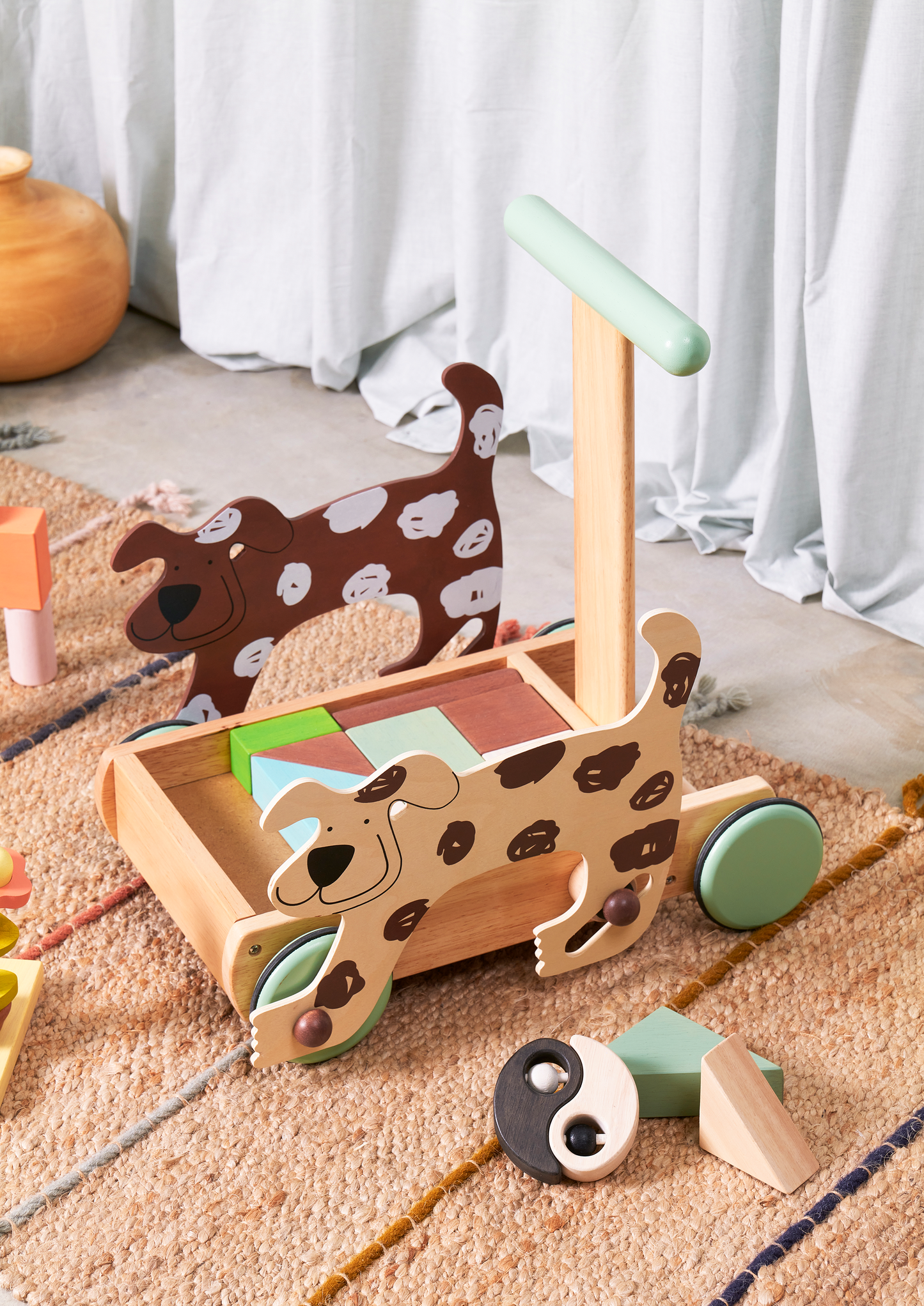 A wooden push toy designed for toddlers, featuring two cartoon-style dogs with spots on each side of the base. The toy has a handle for pushing, four wheels in pastel green, and a colorful geometric pattern on the base. The dogs are smiling, adding a playful and friendly touch.