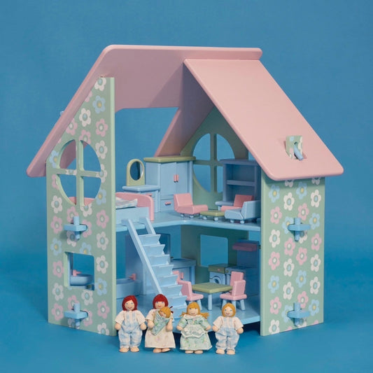 A pastel-colored dollhouse with a pink roof, blue walls, and decorative floral patterns. The dollhouse features a front porch, stairs, and multiple rooms, accompanied by a family of five dolls standing in the front yard.