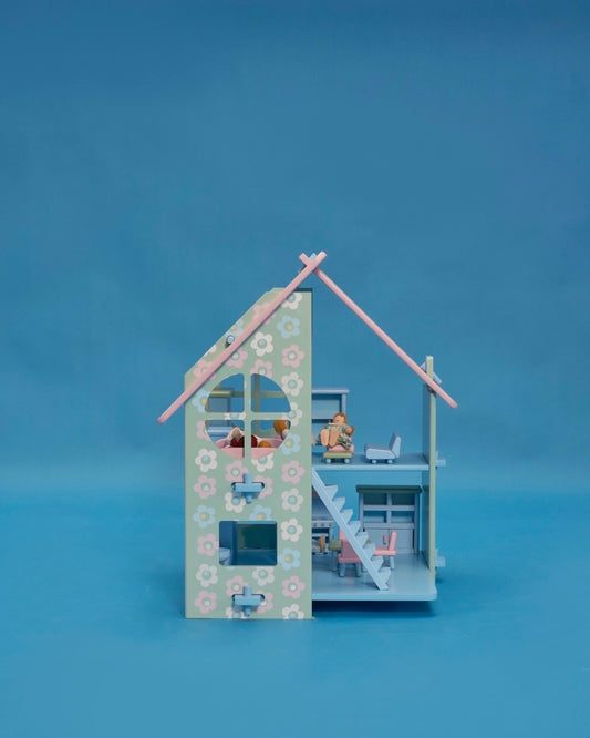 A pastel-colored dollhouse with a pink roof, blue walls, and decorative floral patterns. The dollhouse features a front porch, stairs, and multiple rooms, accompanied by a family of five dolls standing in the front yard.
