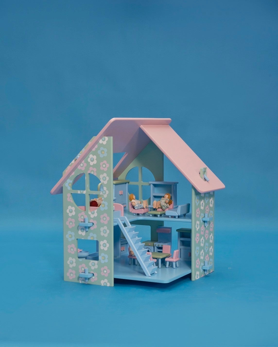 A pastel-colored dollhouse with a pink roof, blue walls, and decorative floral patterns. The dollhouse features a front porch, stairs, and multiple rooms, accompanied by a family of five dolls standing in the front yard.