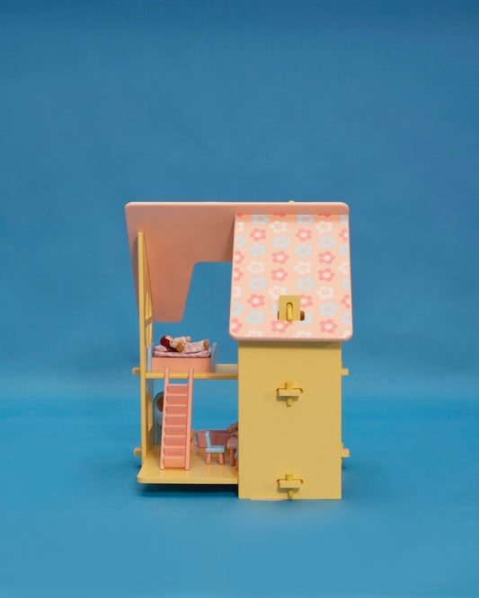A small yellow dollhouse with a pink and patterned roof, featuring a front door, window, and a set of stairs. There are five miniature dolls in the front, dressed in light-colored clothing.