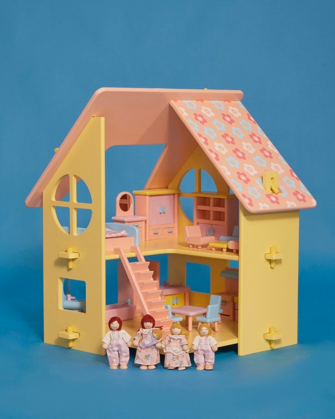 A small yellow dollhouse with a pink and patterned roof, featuring a front door, window, and a set of stairs. There are five miniature dolls in the front, dressed in light-colored clothing.