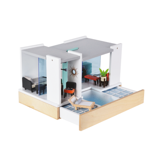 A Mid Century Modern dollhouse with a white and wood facade, featuring a living room with a black sofa, a red armchair, and a blue pendant lamp; a dining area with a wooden table and chairs; and a bedroom with a bed and a wooden headboard.