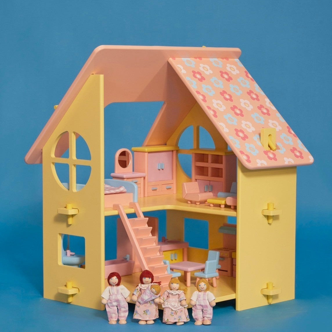 A small yellow dollhouse with a pink and patterned roof, featuring a front door, window, and a set of stairs. There are five miniature dolls in the front, dressed in light-colored clothing.