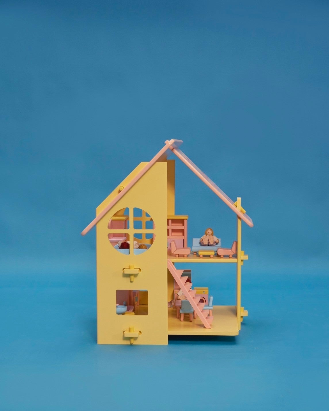 A small yellow dollhouse with a pink and patterned roof, featuring a front door, window, and a set of stairs. There are five miniature dolls in the front, dressed in light-colored clothing.