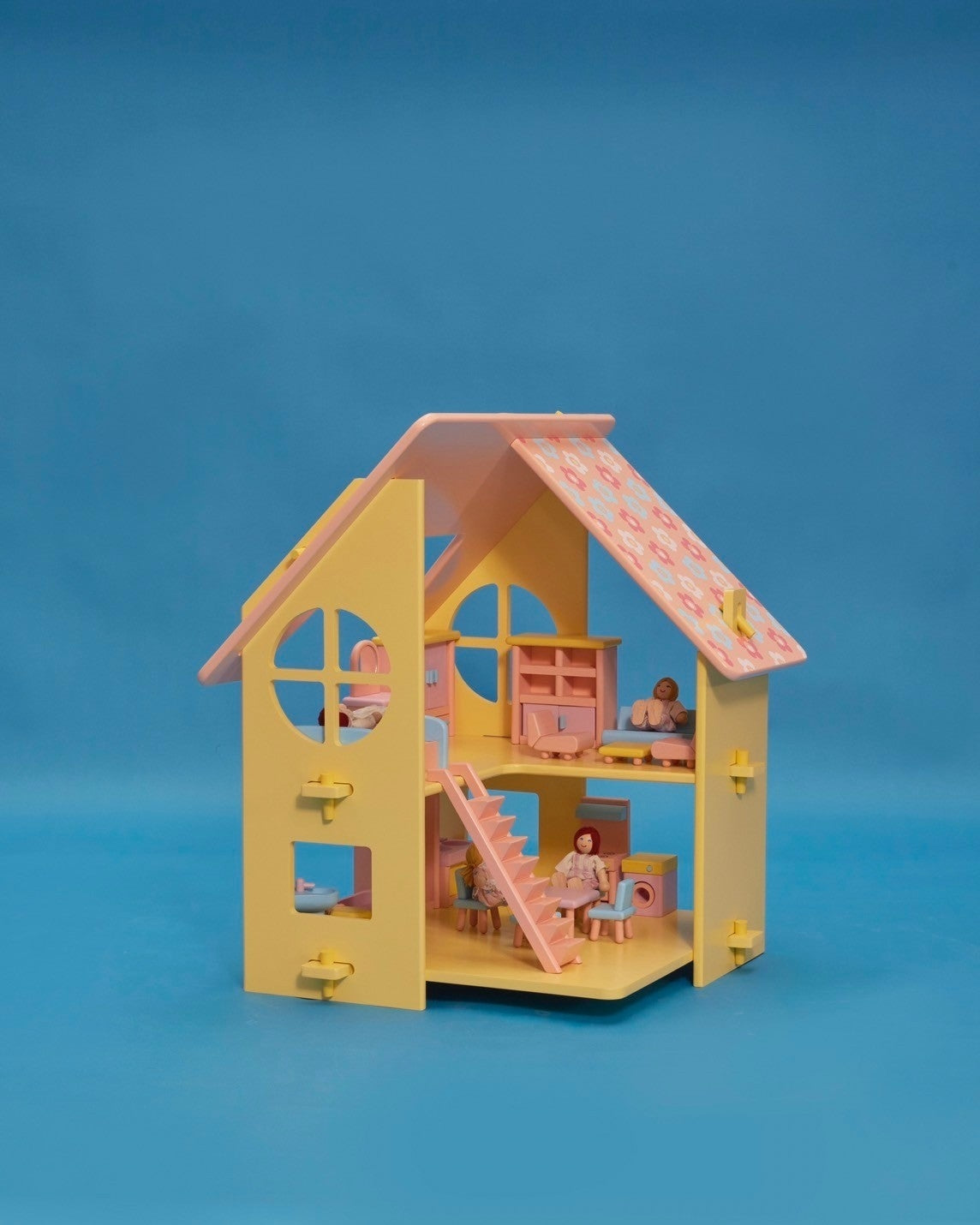 A small yellow dollhouse with a pink and patterned roof, featuring a front door, window, and a set of stairs. There are five miniature dolls in the front, dressed in light-colored clothing.
